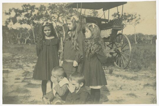 Sepia image of 3 girls standing in front of a buggy and two boys sitting on the ground
