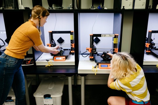 Two people using 3D printers. One person is crouched down examining a creation and the other is standing. 
