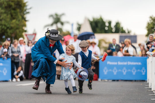 Woman in Edwardian costume and two small children running along street in parade
