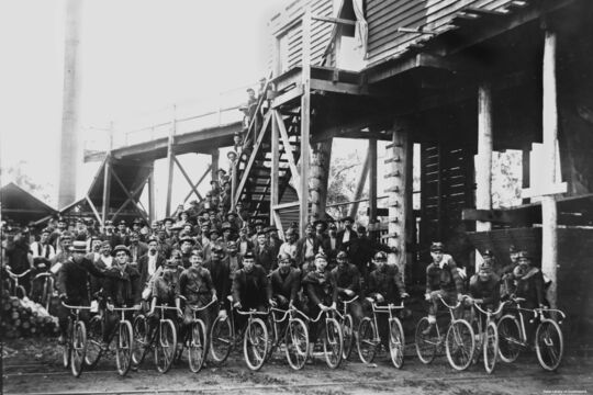 Black-and-white photograph of a large group of miners gathered outside an industrial timber structure, many seated on bicycles in the front row. The men wear work clothes and hats, some with lamps, suggesting a coal mining community. Additional workers stand on stairs and platforms behind them.