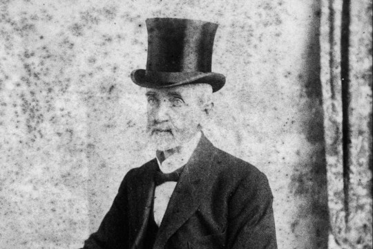 Portrait of man sitting at table wearing top hat and suit with long coat