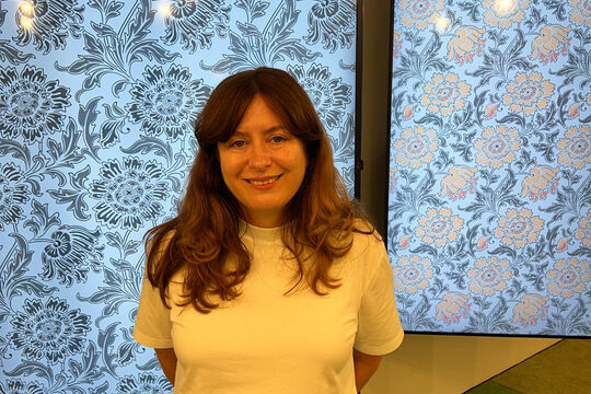Anna Rowe, in a white t-shirt stands in front of two screens that showcase her floral wallpaper work. Her arms are behind her back and she is smiling at the camera, her hair is around her shoulders.