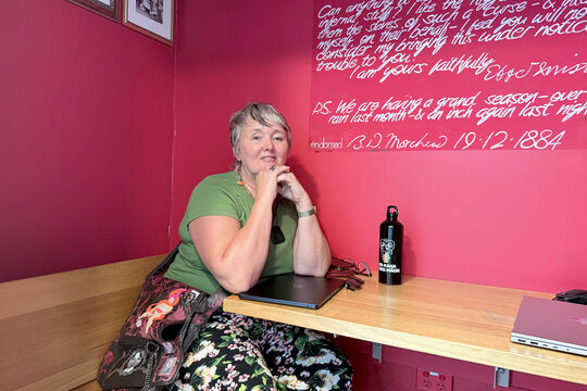 Ruth sits in the Black Opium reading room on Level 4, she is sitting in front of a bright pink wall leaning against the table built into the wall. Her arms are under her chin and her laptop is on the table below her, she is looking at the camera and wearing a green and floral outfit.