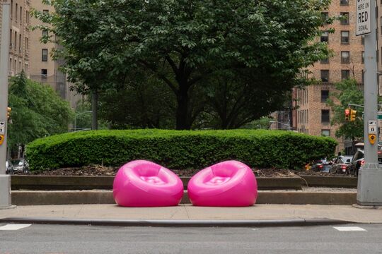 Bright pink inflatable chairs set on a sidewalk in the urban setting of New York, surrounded by neatly trimmed bushes and a tree. Beige buildings and street traffic are visible in the background.