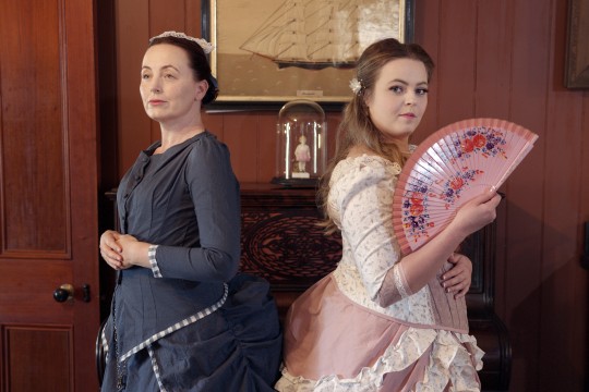 Two female actors wearing colonial dresses, one in dark grey, the other in pink and holding a fan
