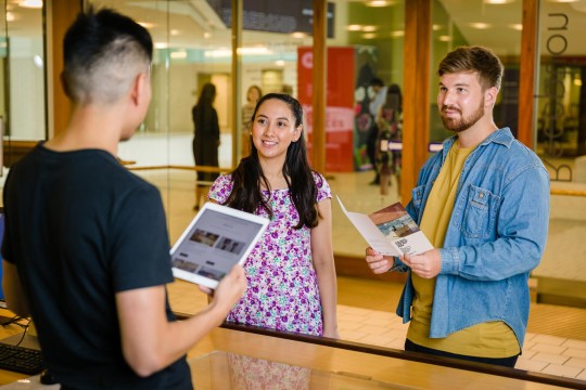 Two visitors talking to a State Library staff member at the reception desk. One visitor is holding a brochure.