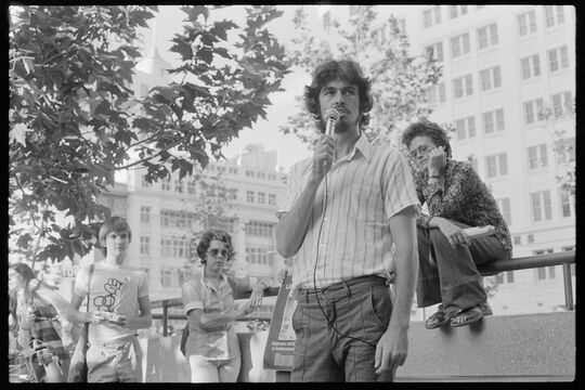 Black and white photograph of a young man speaking into a microphone. Other young people are looking towards him from behind.