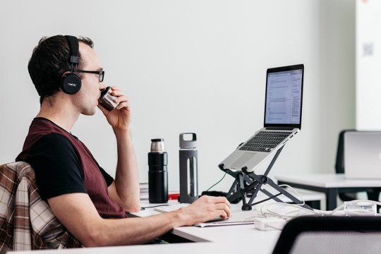 Man sitting at computer with headphones on and drinking coffee