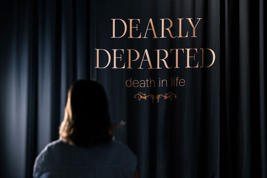 'Dealy Departed' printed on a curtain. A person is seen in the foreground. 
