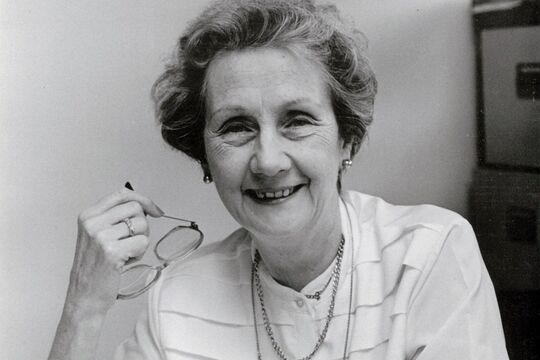 Black-and-white photograph of an older woman smiling at a desk, holding her glasses