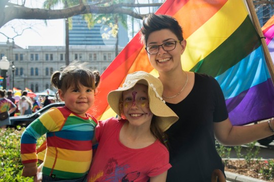 Smiling faces of two children and a person with rainbow clothing and flag.