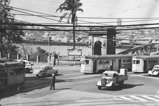 Black-and-white photograph of a busy street scene at the northern end of Victoria Bridge, Brisbane, circa 1960. Trams, cars, and a small truck converge at an intersection while a uniformed traffic officer stands in the roadway directing vehicles.