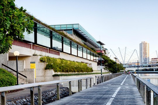 View of the exterior of the State Library building from the Brisbane River with Kurilpa Bridge and buildings in the background. 