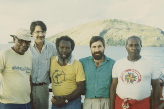 5 men stand on a boat in front of an island