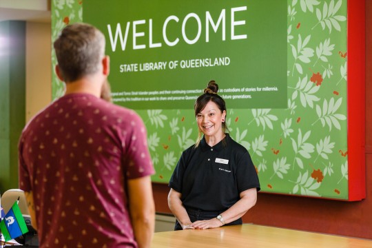 Client facing a smiling State Library staff member at the reception desk. The wall behind the staff member says 'Welcome'.