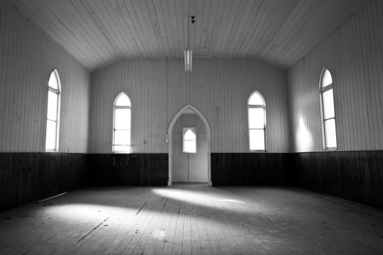 The inside of an empty wood paneled church. Sunlight streams through big arched windows.