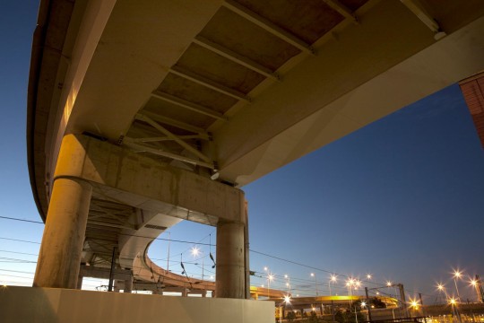 The underside of a Brisbane overpass, at dusk with the street lights glowing yellow in the background.