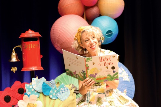 A picture of a woman in bright clothing on stage smiling at audience and holding a book "hotel for bees'