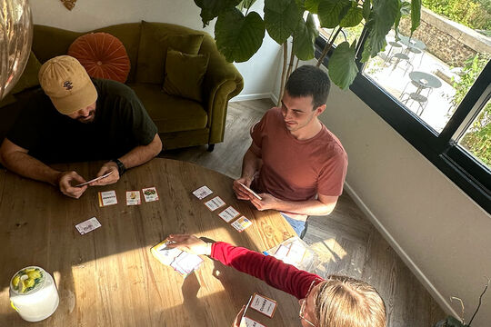 Three people sitting around a light coloured round table playing the cooked game, three people are playing and there are a bunch of cards in the table with each person holding a bunch of cards in their hands. 
