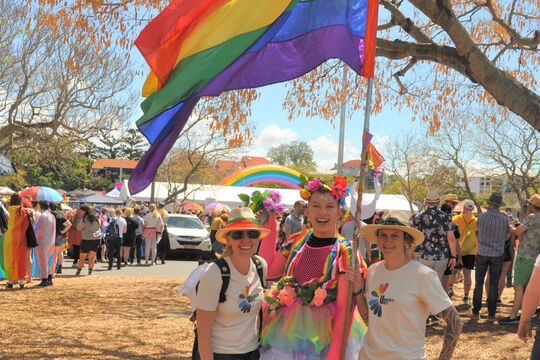 Three women standing together in a park holding a rainbow flag with crowd in background
