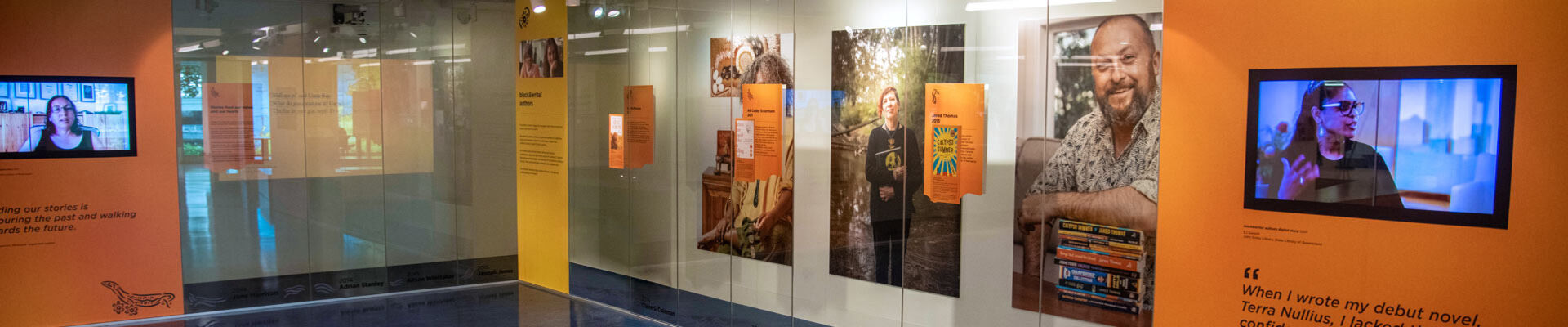 Inside the Sovereign Stories showcase at State Library. Large portraits of featured authors and publishers are on the wall behind glass and two screens are playing recorded interviews.