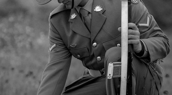 Black and white photo of a solider kneeling tp pick a poppy from a grassy field. Poppy flowers have been coloured red and are the only colour in the photo. 