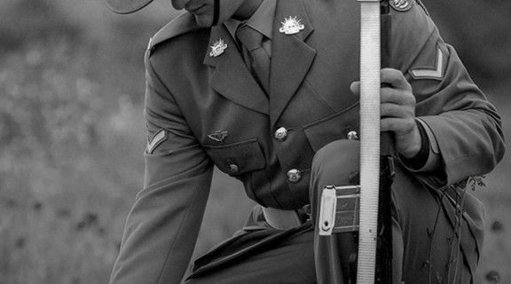 Black and white photo of a solider kneeling tp pick a poppy from a grassy field. Poppy flowers have been coloured red and are the only colour in the photo.