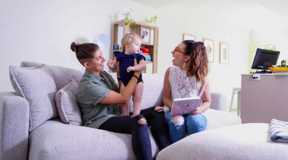 Two people sitting on a couch at home with a book. One person has a young child on her knee.