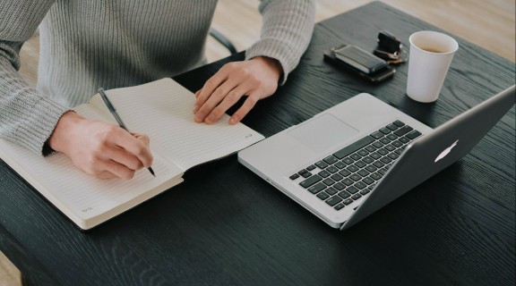 A person is writing noted in a note book. A laptop, coffee cup, wallet and keys is also on their desk.