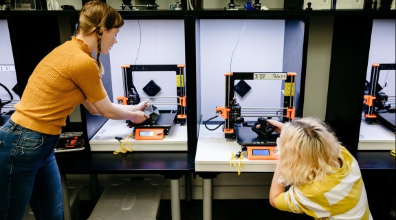 Two people using 3D printers. One person is crouched down examining a creation and the other is standing.
