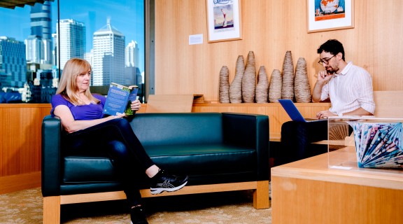 Two people sitting inside at the library. One person is reading a book and another is using a laptop.