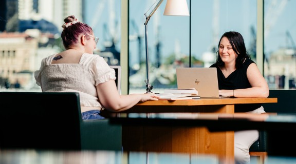 Two women sitting at a joint desk with a view of Brisbane city using laptops at State Library.