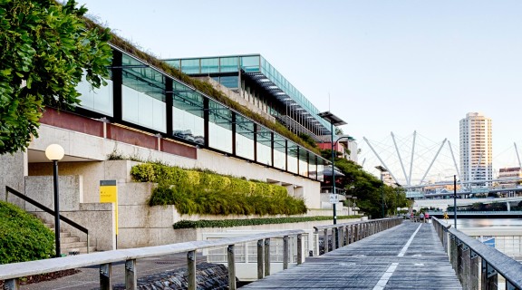 View of the exterior of the State Library building from the Brisbane River with Kurilpa Bridge and buildings in the background.