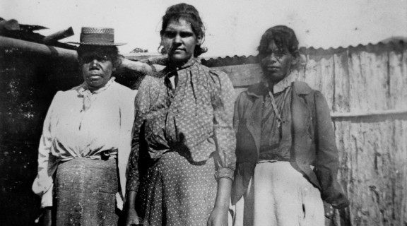 Three First Nations women standing in front of a wooden shed