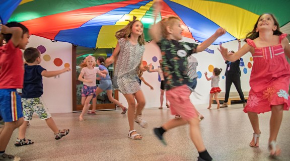 a group of children smiling and running under a colourful parachute