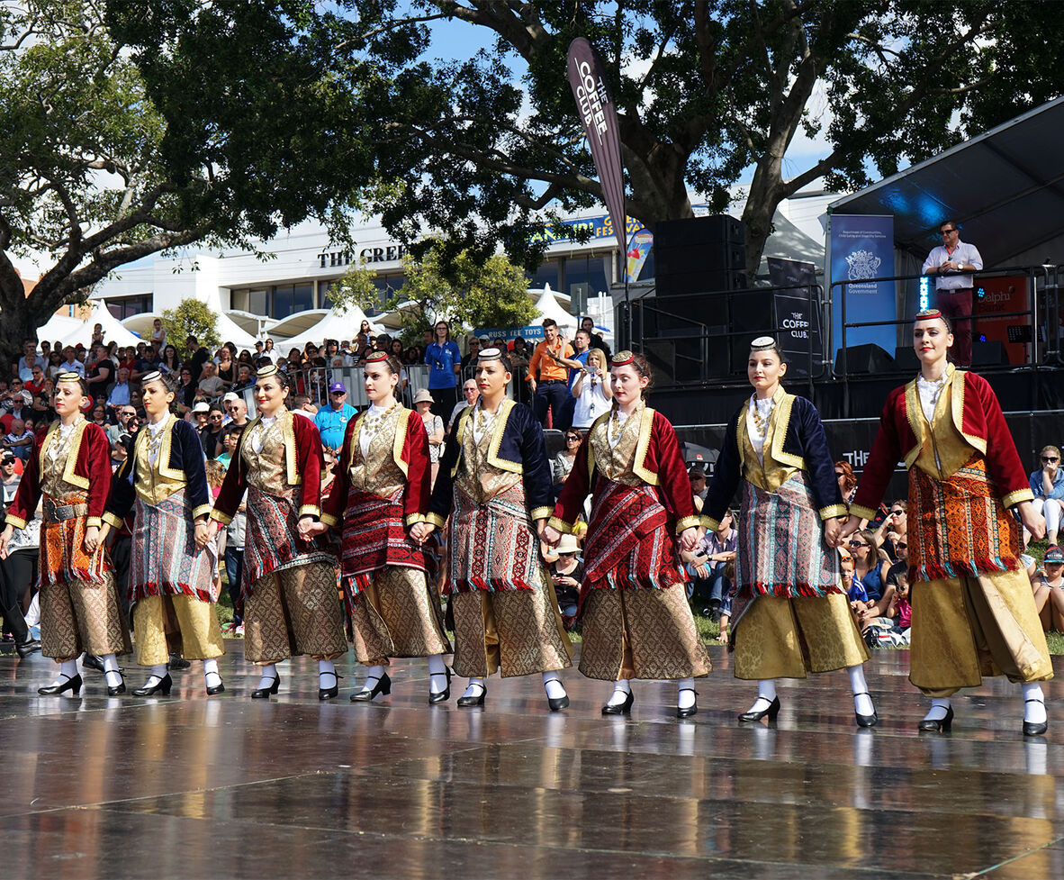 Women dressed in traditional Greek clothing preforming a dance on stage.