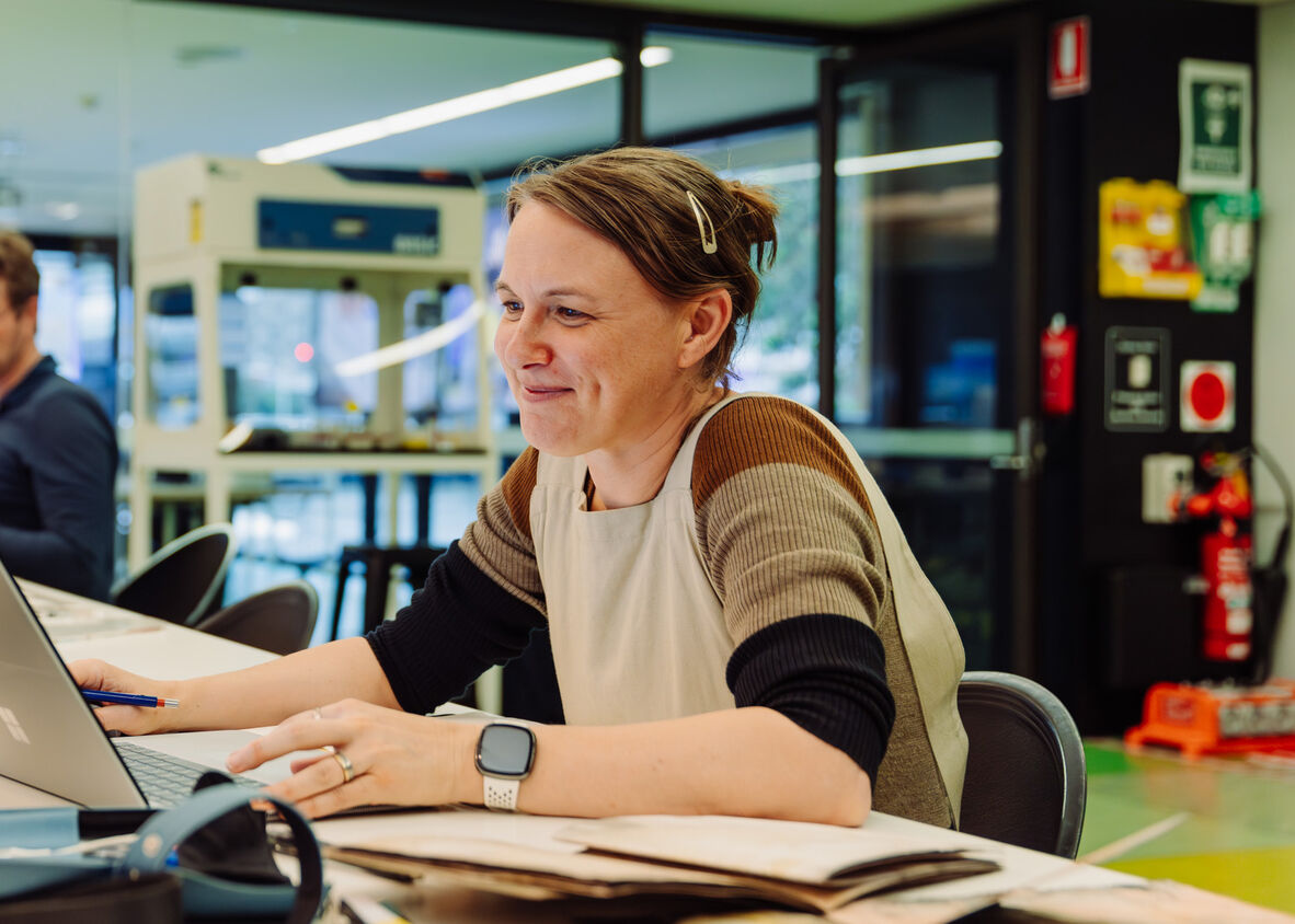 A photo of a woman at a desk working at a laptop with papers and books around her
