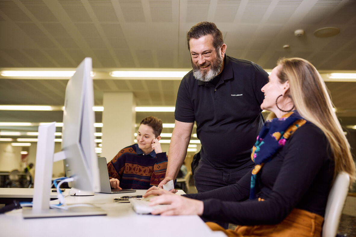 A man assists a woman at a computer, while another person works on a laptop in the background.