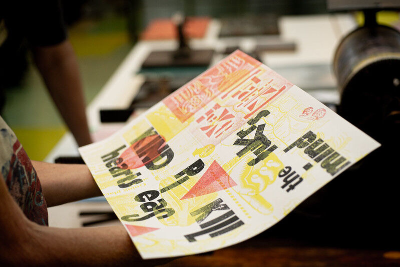 Person holds an A3 sheet with yellow, red and black printing on it with words and shapes