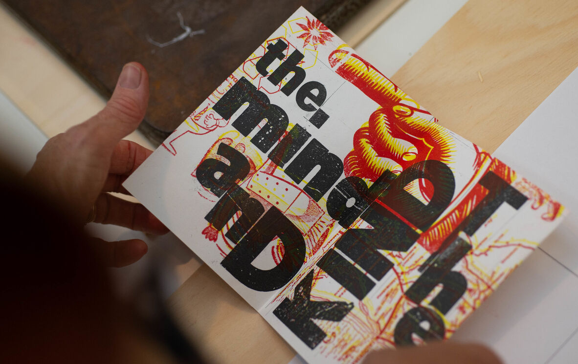 a person holds their artist book with black writing and red and yellow prints of hands and maps 
