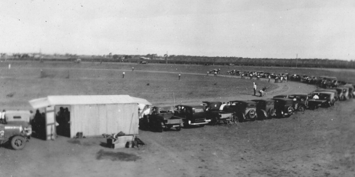 Black-and-white photograph of a bicycle race at a rural track, with cyclists rounding a distant curve. A long line of early motor cars is parked beside the course, and spectators stand nearby. A small shed sits in the foreground, with open fields and trees in background.