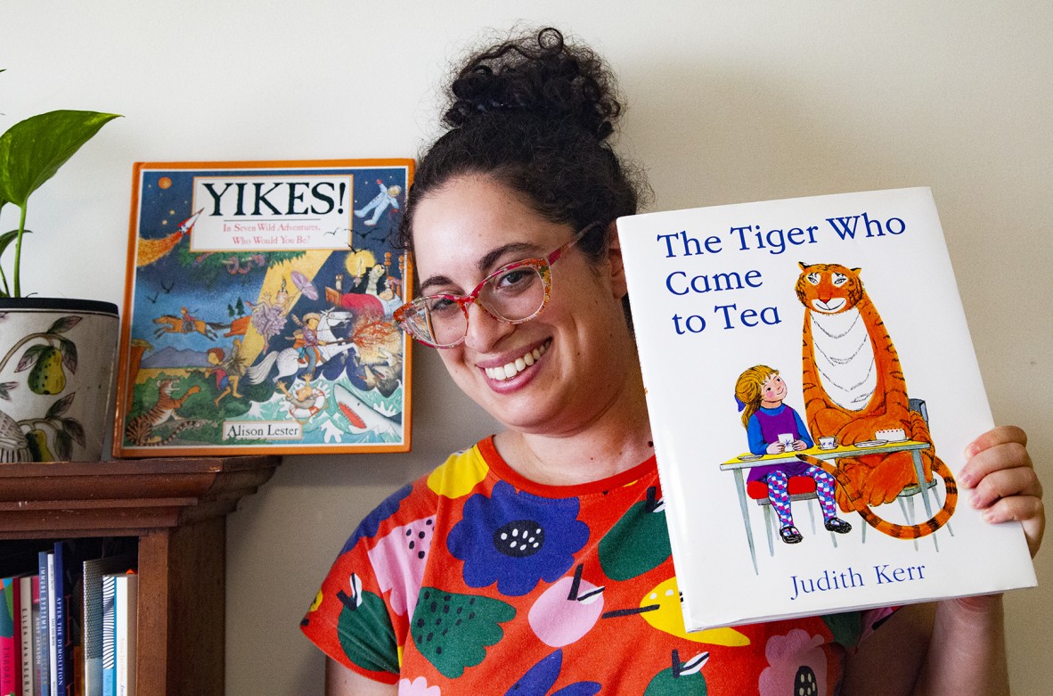 Anna Jacobson stands beside a bookshelf, wearing a bright floral shirt and holding up a copy of 'The tiger who came to tea'
