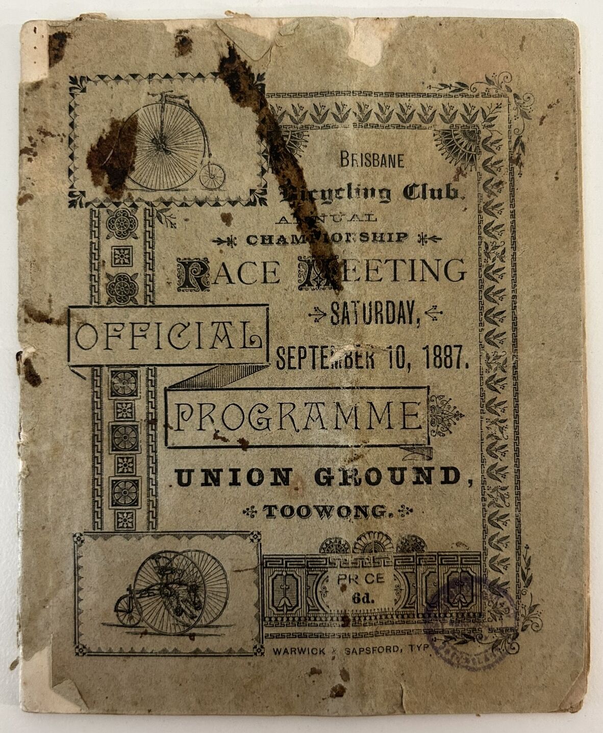 Worn and stained printed programme for the Brisbane Cycling Club Annual Championship Race Meeting, held Saturday, September 10, 1887, at Union Ground, Toowong. Decorative borders and illustrations of early bicycles frame the text, with visible creases, tears, and age-related damage across the paper.