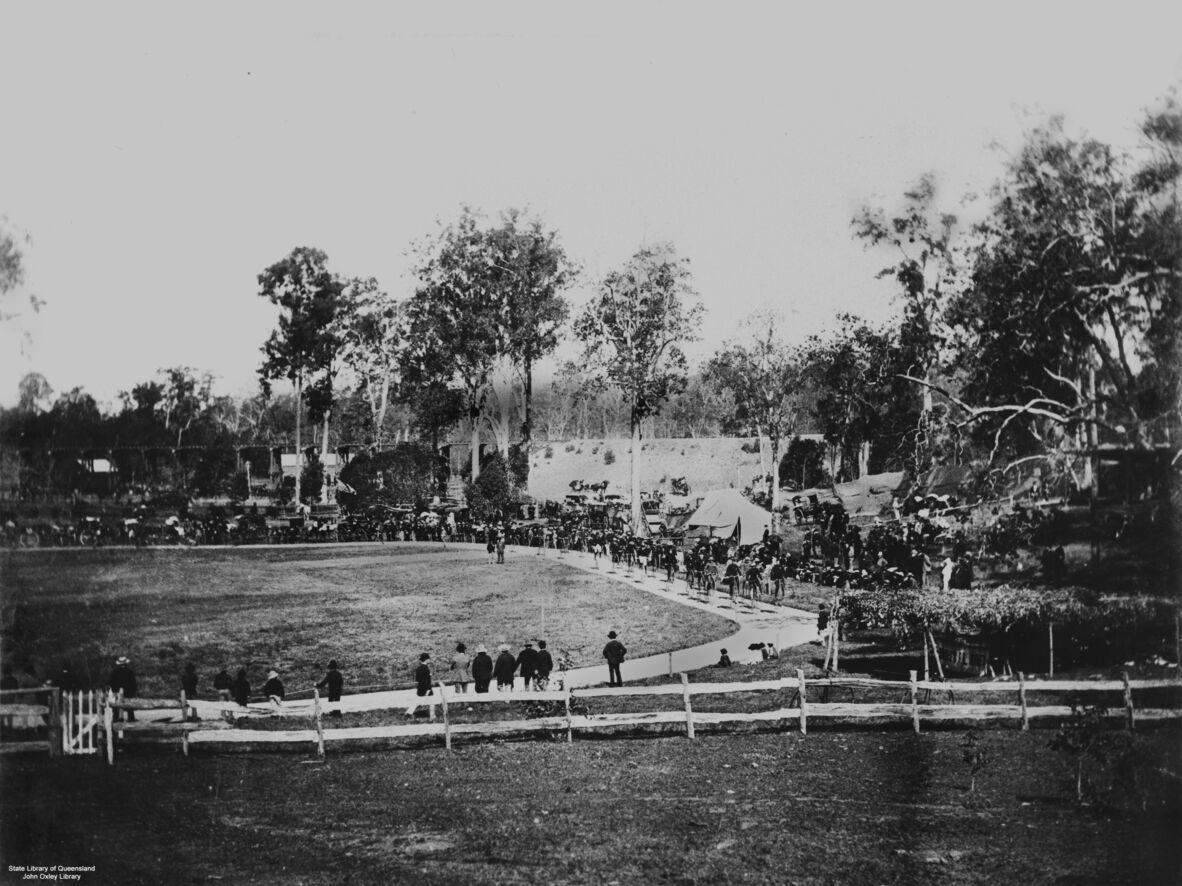 Black-and-white photograph of a bicycle racing event at Toowong, Brisbane, circa 1885. Cyclists ride along a curved track bordered by spectators, tents, and horse-drawn carriages. Large trees surround the grounds, while crowds gather along pathways and fences to watch the race.