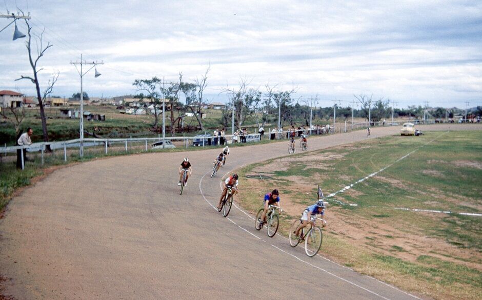Colour photograph of cyclists racing around a dirt velodrome at Miles End, Mount Isa, circa 1956. Riders lean into a curved track while spaced in a line. Spectators stand along a fence, with scattered buildings, trees, and parked cars visible under a cloudy sky.