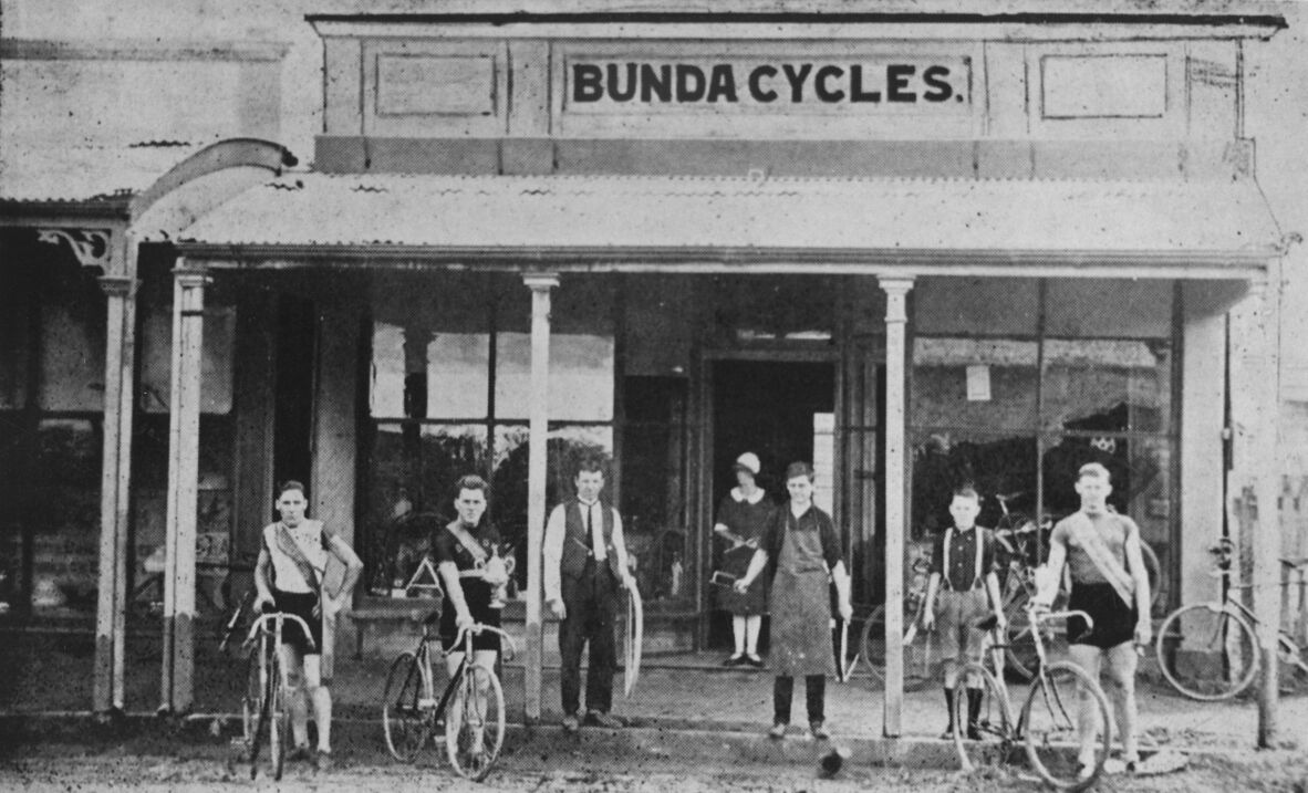 Black-and-white photograph of the Bunda Cycles shopfront in Bundaberg, circa 1920s. Several young men and boys stand with bicycles outside the timber building, while a woman stands in the doorway. Large windows display cycles inside, and a sign reading “Bunda Cycles” is mounted above.