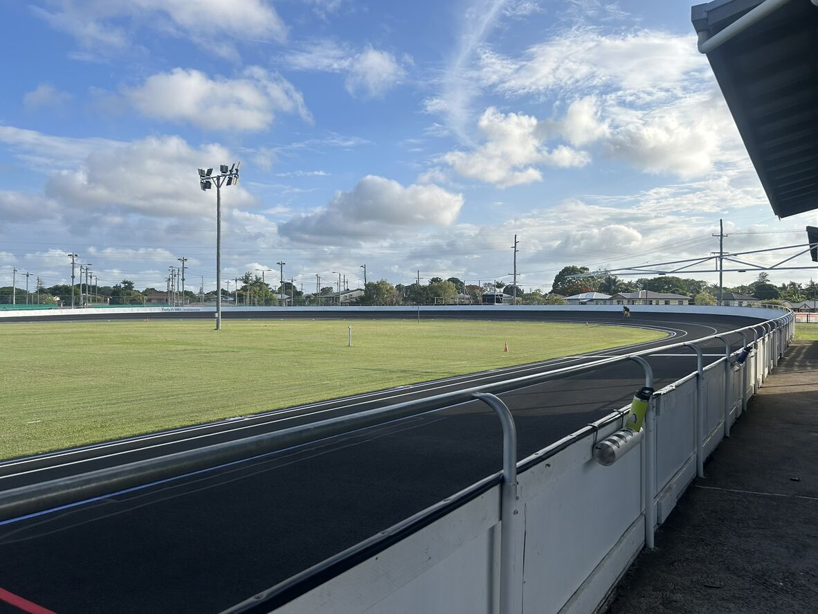 Wide view of a modern outdoor velodrome with a smooth black track curving around a grassy infield. A lone cyclist rides along the far bend. Metal railings line the foreground, with floodlights, houses, and trees beneath a partly cloudy blue sky.
