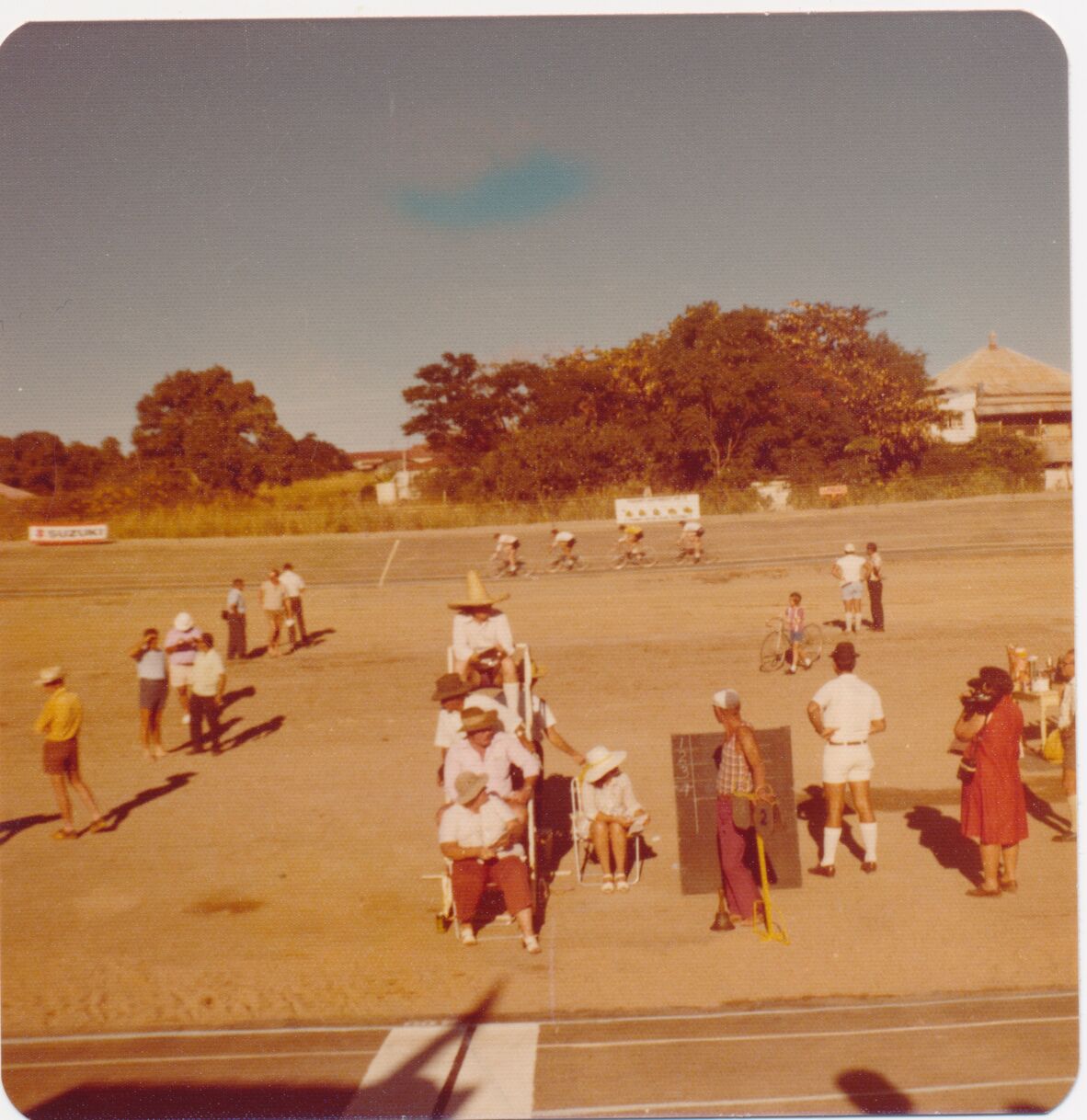 Faded colour photograph of a velodrome cycling race, with riders circling the track in the background. Spectators and officials stand and sit in the foreground, some wearing hats. A scoreboard and signage are visible, with trees and buildings beyond, suggesting a warm, outdoor sporting event setting.