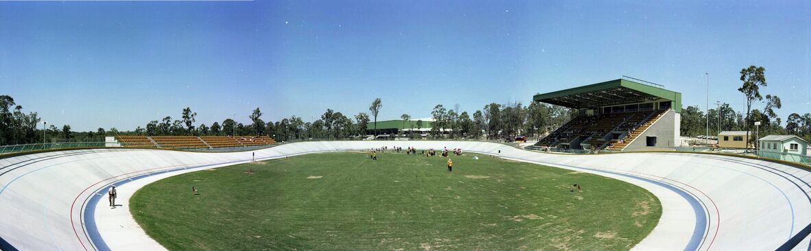 Wide panoramic colour photograph of a modern velodrome at Chandler, featuring a smooth concrete track surrounding a grassy infield. Tiered seating and a covered grandstand overlook the arena, with scattered people on the field. Trees and buildings frame the venue beneath a clear blue sky.