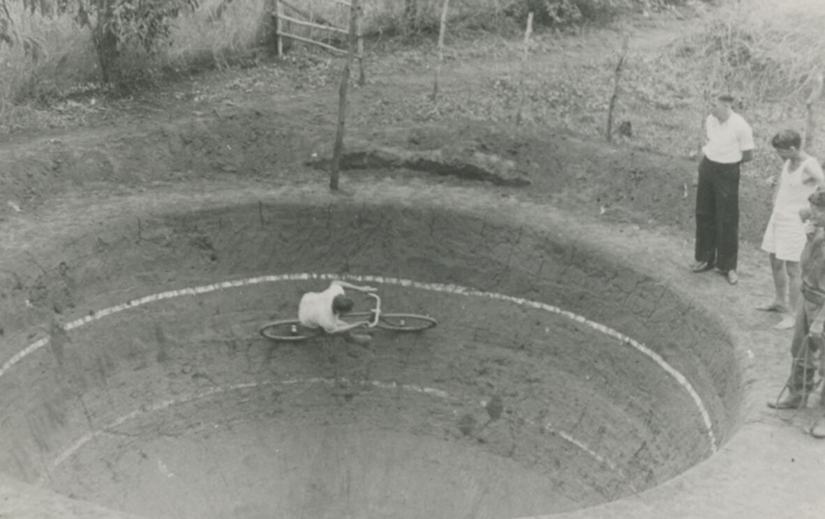 Black-and-white photograph of a cyclist riding low along the inside wall of a hand-dug dirt velodrome. The steep circular track forms a bowl shape, with a few onlookers standing at the rim. Trees, fencing, and rough ground surround the unusual, improvised cycling venue.